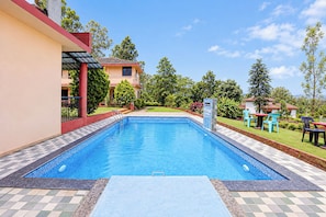 Una piscina con borde de baldosas, rodeada de vegetación y un edificio con tejado rojo.