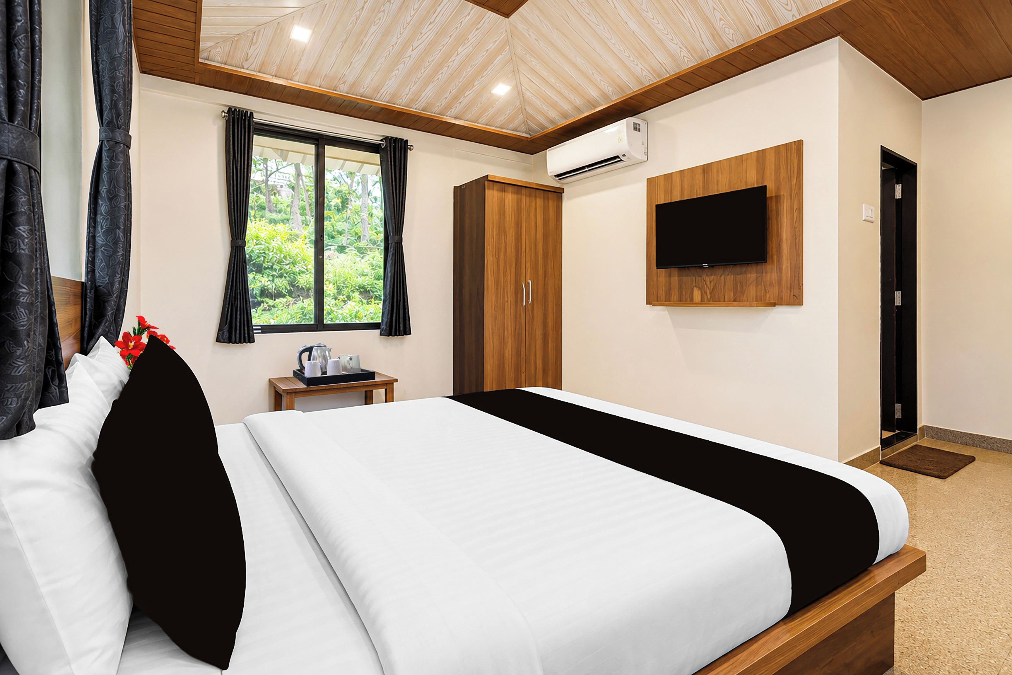 A hotel room with a bed, a television mounted on the wall, a window with curtains, and an air conditioning unit.