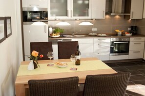 Une cuisine moderne avec des armoires blanches, des appareils en acier inoxydable et un coin repas doté de chaises en osier et d’une table dressée pour deux personnes.