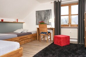 Ein Schlafzimmer mit zwei Betten, einem Schreibtisch, einem Stuhl, einem roten Hocker, einem Fenster mit Vorhängen und einem Bild an der Wand.