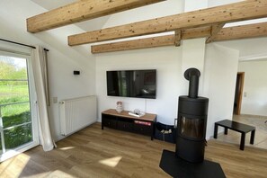 A modern living room with a large flat-screen TV, a wood-burning stove, and a sliding glass door to a garden. - Country House Maarweg (Loquard)