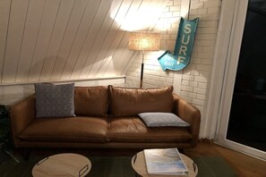A brown leather sofa with two cushions, a small wooden table with papers, a lamp, and a 'SURF THIS WAY' sign on a brick wall. - Holiday home Burhave (Burhave)