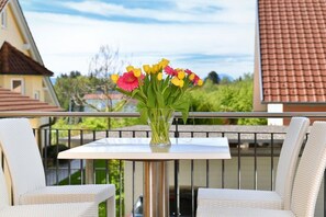 A balcony with a table and two wicker chairs, a vase with flowers, and a view of houses and greenery. - C - Dinkelbach Object 1 (Lindau)