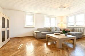 Ein Wohnzimmer mit einer Couch, einem Couchtisch und einer Lampe. Es gibt Fenster und eine Heizung.