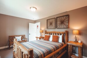 A bedroom with a wooden bed, a bench, two wall-mounted lamps, and a door with a fire extinguisher. - Hidden Meadows | Hocking Hills, Sleeps 14, Hot Tub (Rockbridge)