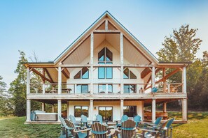 A two-story wooden house with a balcony, outdoor seating, and a large window. - Hidden Meadows | Hocking Hills, Sleeps 14, Hot Tub (Rockbridge)