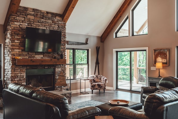 A spacious living room with a stone fireplace, leather sofas, and large windows. - Hidden Meadows | Hocking Hills, Sleeps 14, Hot Tub (Rockbridge)