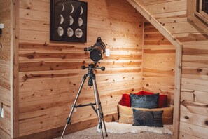 A wooden cabin interior with a telescope on a tripod, a framed picture of the moon phases, and a cozy seating area with cushions. - Hidden Meadows | Hocking Hills, Sleeps 14, Hot Tub (Rockbridge)