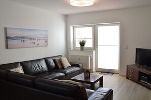 A modern living room with a black leather sofa, a coffee table, a flat-screen TV, and a large window with a view of the outdoors. - Holiday apartment with 2 bedrooms (Borkum)