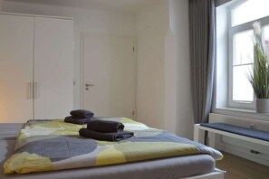 Ein Schlafzimmer mit einem Bett, einer Bank, einer Zimmerpflanze und einem Fenster mit Vorhängen.