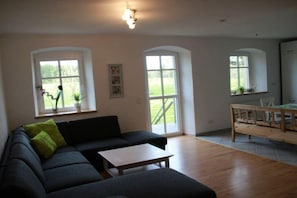 Ein Wohnzimmer mit einem dunklen Sofa, einem Holztisch und einer Holzbank. Drei Fenster sorgen für natürliches Licht.