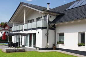 Une maison moderne à deux étages, dotée d’un balcon, de grandes fenêtres et d’un espace extérieur aménagé et abrité.