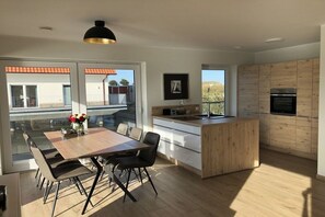 A modern kitchen with wooden cabinets, a central island, and a dining area with a table and chairs. - Pearl at the marina, Pfahlhaus Harlesiel (Carolinensiel-Harlesiel)