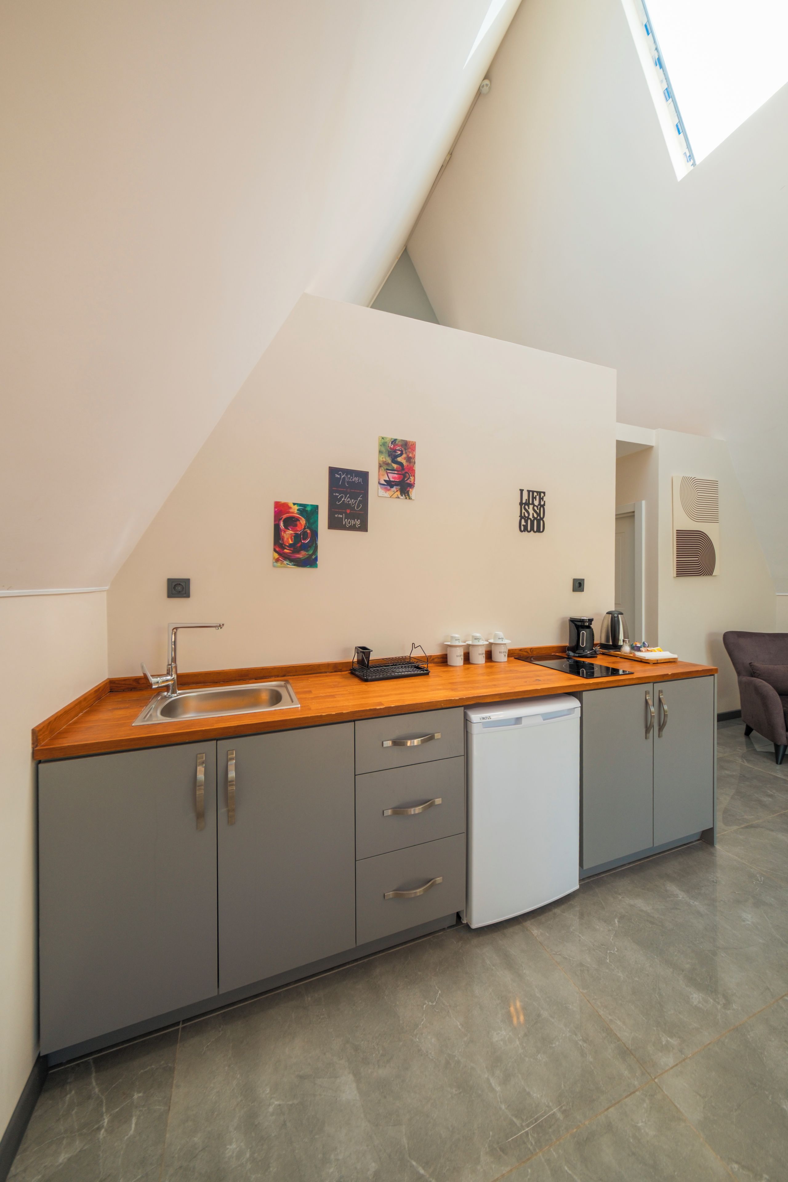 A modern kitchen with grey cabinets, a wooden countertop, a dishwasher, and a coffee maker. There are three wall art pieces and a motivational quote on the wall.