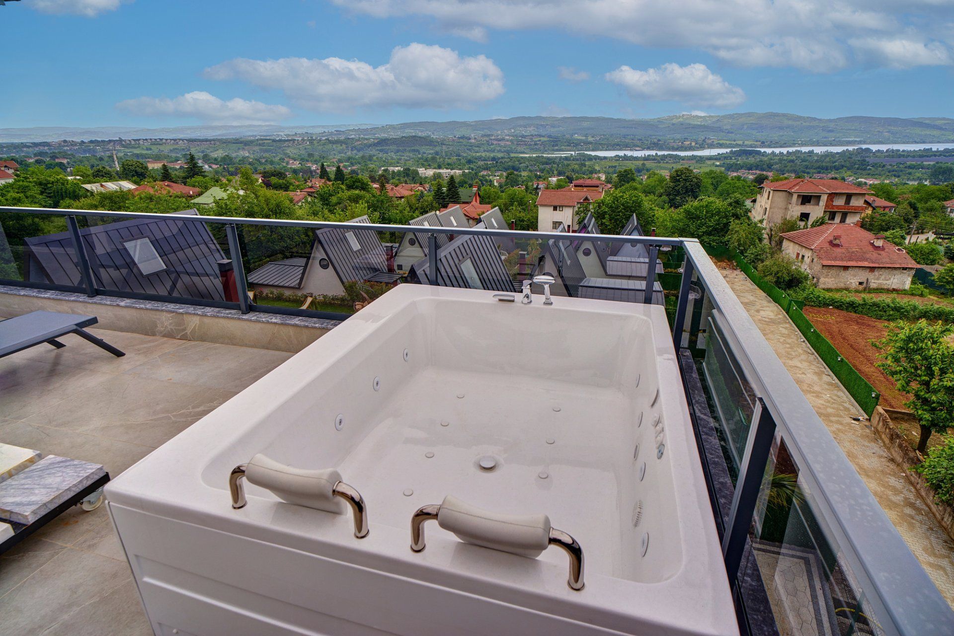 A rooftop terrace with a hot tub, a lounge chair, and a view of a hilly landscape.