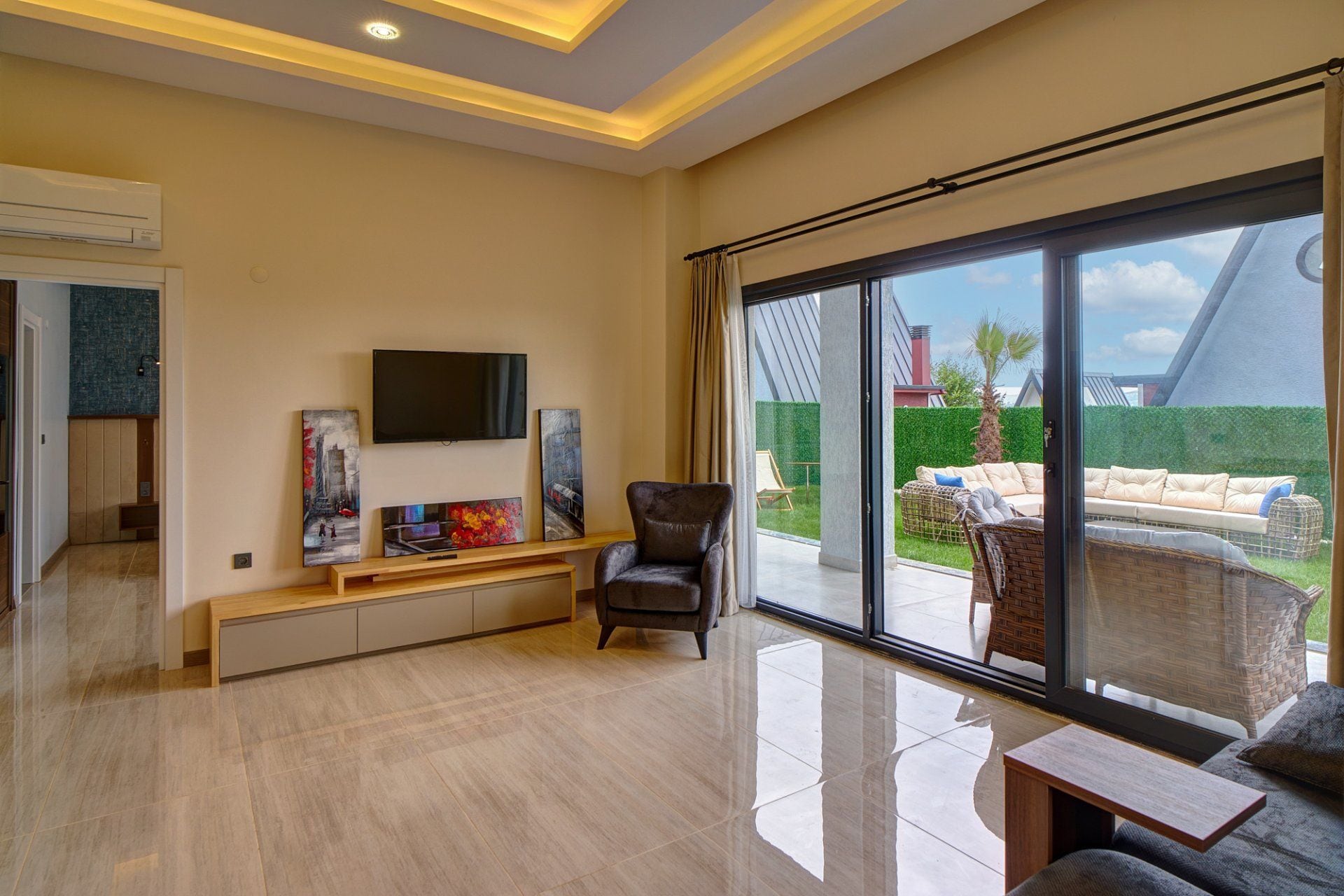 Amplia sala de estar con televisor de pantalla plana, obras de arte, un sillón cómodo, un sofá y un gran ventanal corredero que da a un espacio exterior.