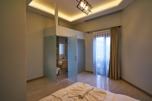 A modern hotel room with a glass-enclosed shower, a large bed with a white comforter, and a view of the outdoors through a large window with curtains.