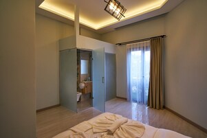 Habitación de hotel moderna con ducha envuelta en vidrio, una cama grande con edredón blanco y vistas al exterior a través de un ventanal con cortinas.