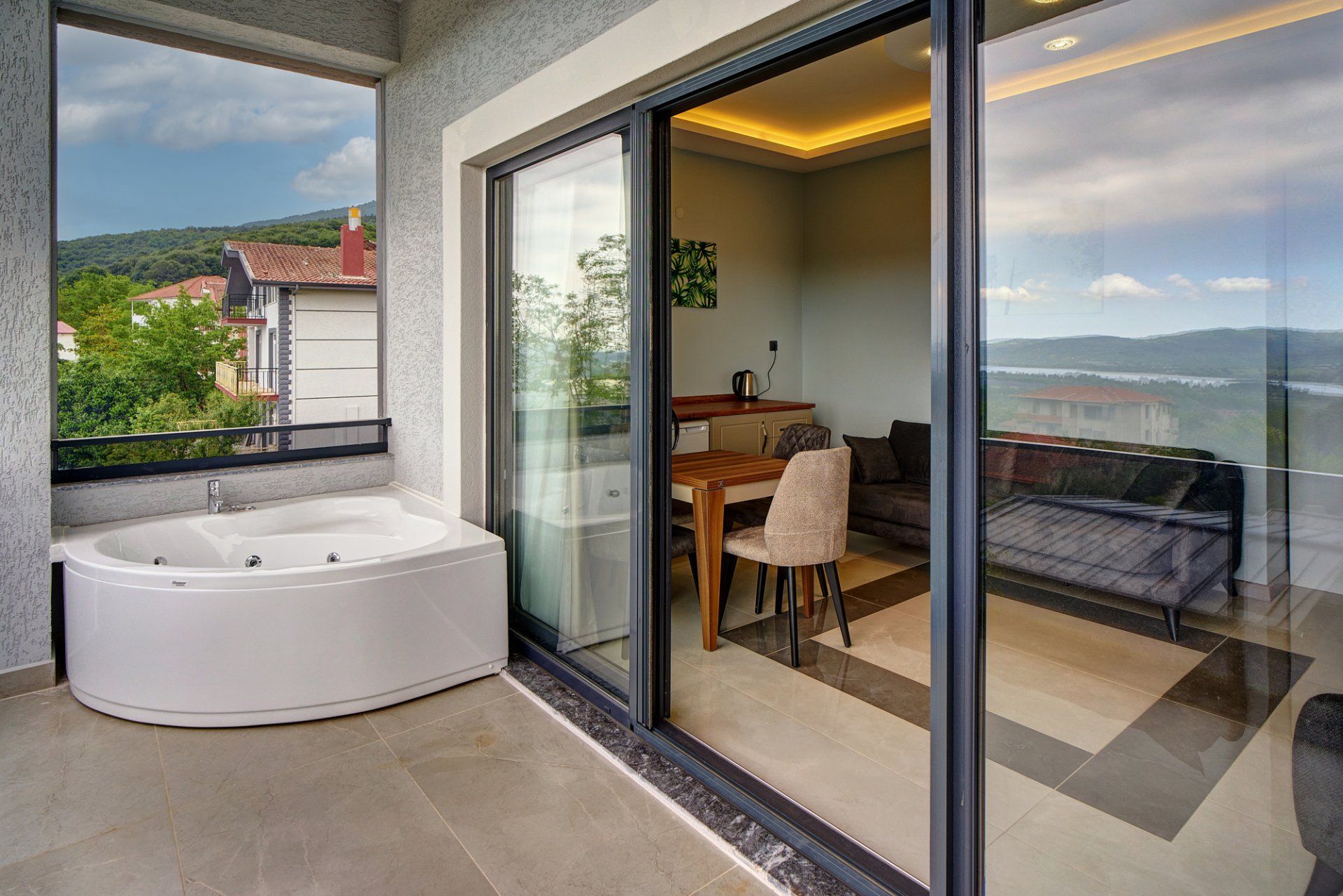 A modern bathroom with a freestanding bathtub, a glass-enclosed balcony with a dining area, and a view of a mountainous landscape.