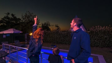 Un grupo de personas de pie junto a una piscina con barandilla, con vistas a un jardín arbolado y un cielo despejado.