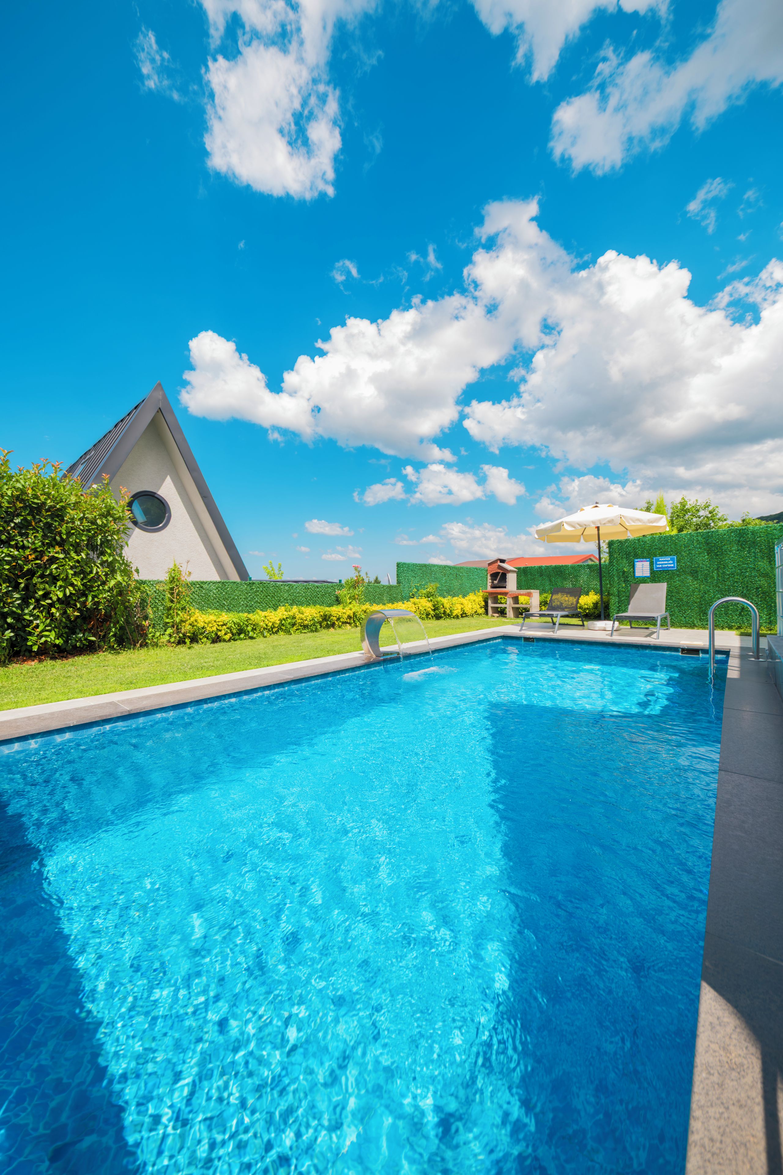 Una piscina con agua azul cristalina, rodeada de un jardín cuidado y un edificio con un techo triangular distintivo.