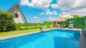 Una piscina con agua azul cristalina, rodeada de un jardín cuidado y un edificio con un techo triangular distintivo.