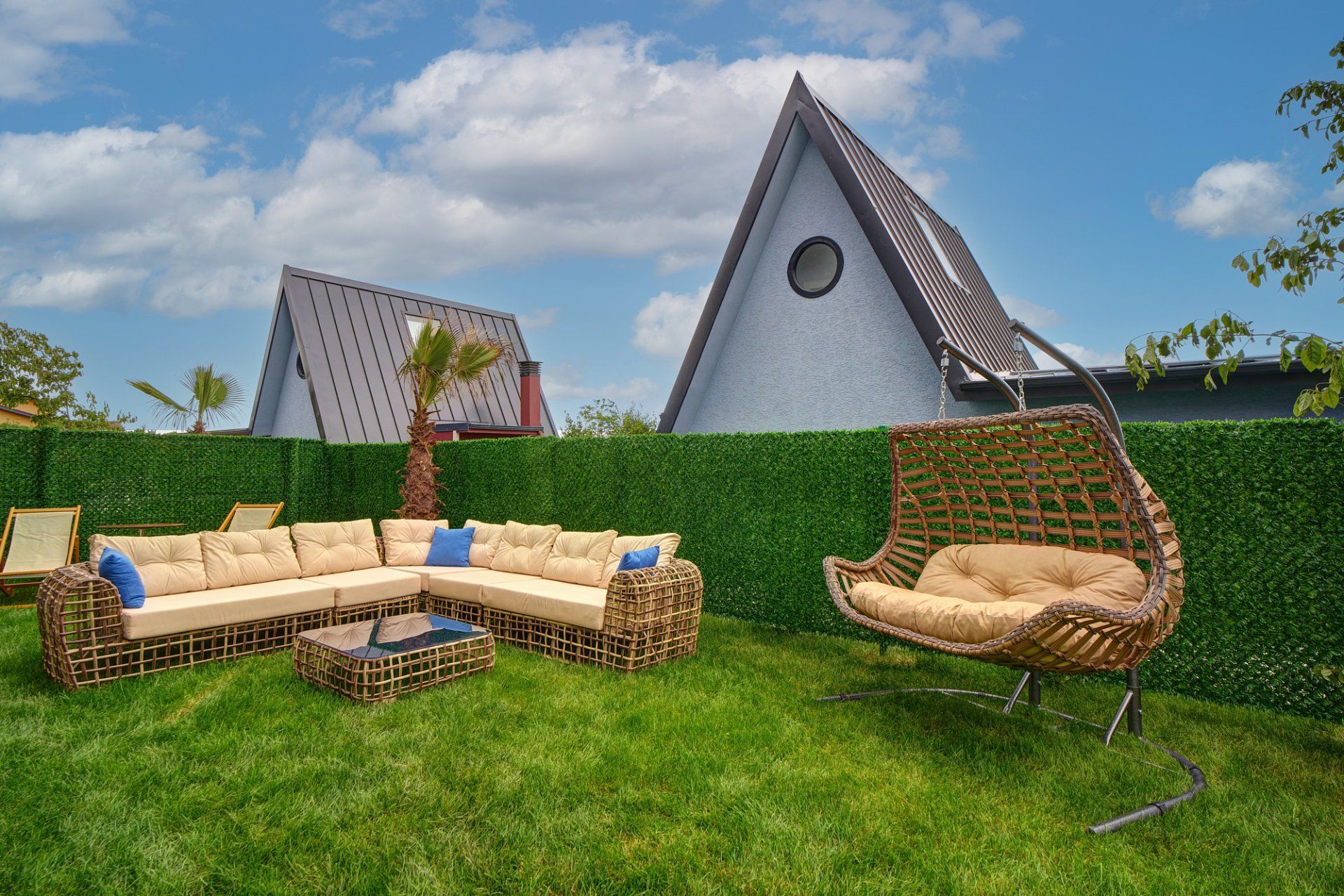 A modern outdoor seating area with a sofa and a wicker chair, a neatly trimmed hedge, and a unique triangular building in the background.