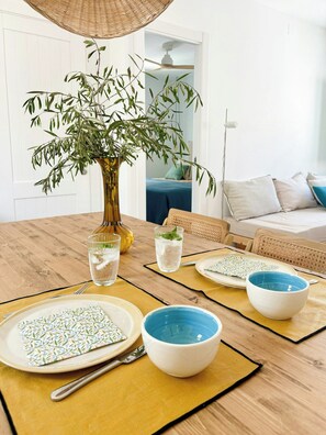 Une table à manger dressée avec des assiettes, des bols et des verres, un vase avec une plante et un luminaire suspendu en osier.