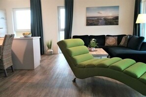 A modern living room with a green sofa, a white coffee table, and a black couch. There is a window with curtains, a painting on the wall, and a plant on the coffee table. - Comfortable apartment with panoramic views (Wangerland-Schillig)