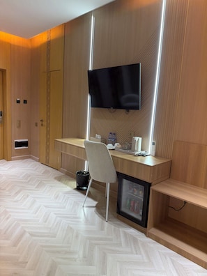 Una habitación de hotel moderna con suelo de madera, un escritorio con silla, un televisor montado en la pared y un refrigerador pequeño.