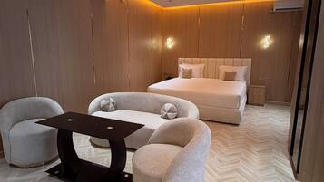 A modern bedroom with a bed, two armchairs, a small table, and a herringbone-patterned floor.