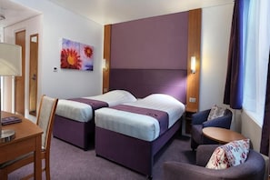 Ein Hotelzimmer mit zwei Betten, einem Schreibtisch, einem Stuhl, einem kleinen Tisch und einem Fenster mit Vorhängen.