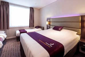 Ein Hotelzimmer mit zwei Betten, einem großen Fenster mit Vorhängen, einem Bettkopfteil mit integrierter Beleuchtung und einer Bettdecke mit einem Logo.