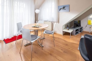 Ein modernes Wohnzimmer mit Essbereich, ausgestattet mit einem runden Tisch, Stühlen, einem roten Teppich, einem Fernseher und einem Bücherregal.