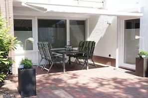 An outdoor patio with a table and chairs, a potted plant, and a window with blinds. - Holiday apartment with 1 bedroom (Bad Zwischenahn)