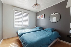 Quarto com duas camas, um radiador, janela com persianas e uma parede com duas obras de arte emolduradas.