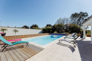 Una piscina con sillones de descanso, una terraza de madera y una pared blanca.