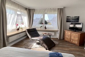 A modern bedroom with a large window, a leather chair, a bed, a TV, and a wooden dresser. - Holiday apartment with 2 bedrooms (Westerland\/Sylt)