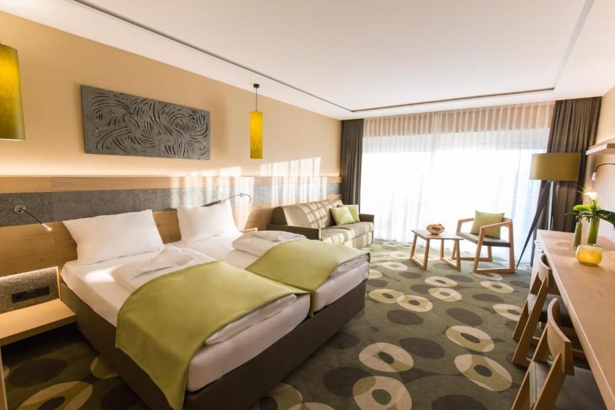 Habitación de hotel con una cama grande, un sofá, un escritorio y una silla. Hay una ventana con cortinas, un cuadro en la pared y una lámpara.