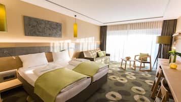 Ein Hotelzimmer mit einem großen Bett, einem Sofa, einem Schreibtisch und einem Stuhl. Ein Fenster mit Vorhängen, ein Bild an der Wand und eine Lampe sind ebenfalls vorhanden.