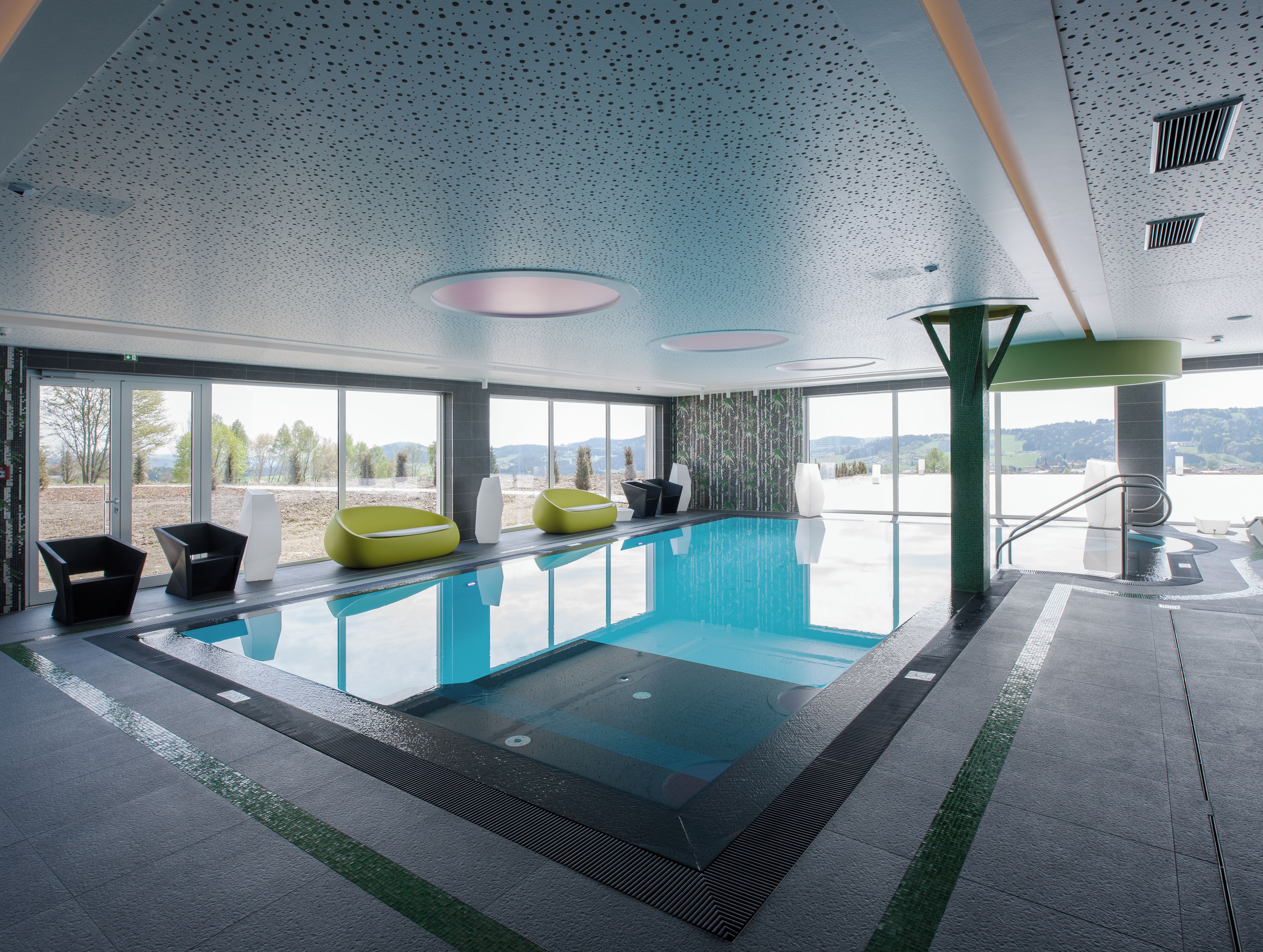 Piscina interior moderna con paredes de vidrio transparente, vistas al exterior y mobiliario contemporáneo.