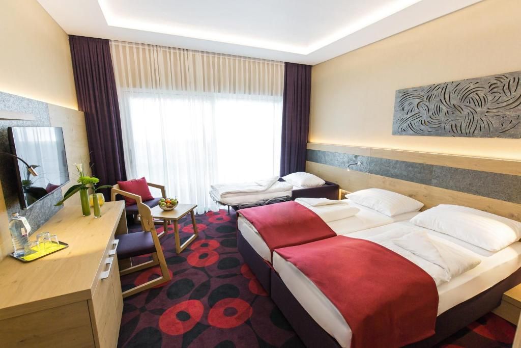 Habitación de hotel con cama, escritorio, silla y un ventanal con cortinas.