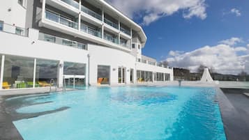 A modern hotel with a large outdoor swimming pool, multiple balconies, and a clear sky.
