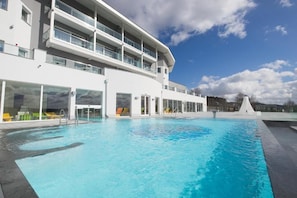 Un hotel moderno con una amplia piscina exterior, varios balcones y cielo despejado.