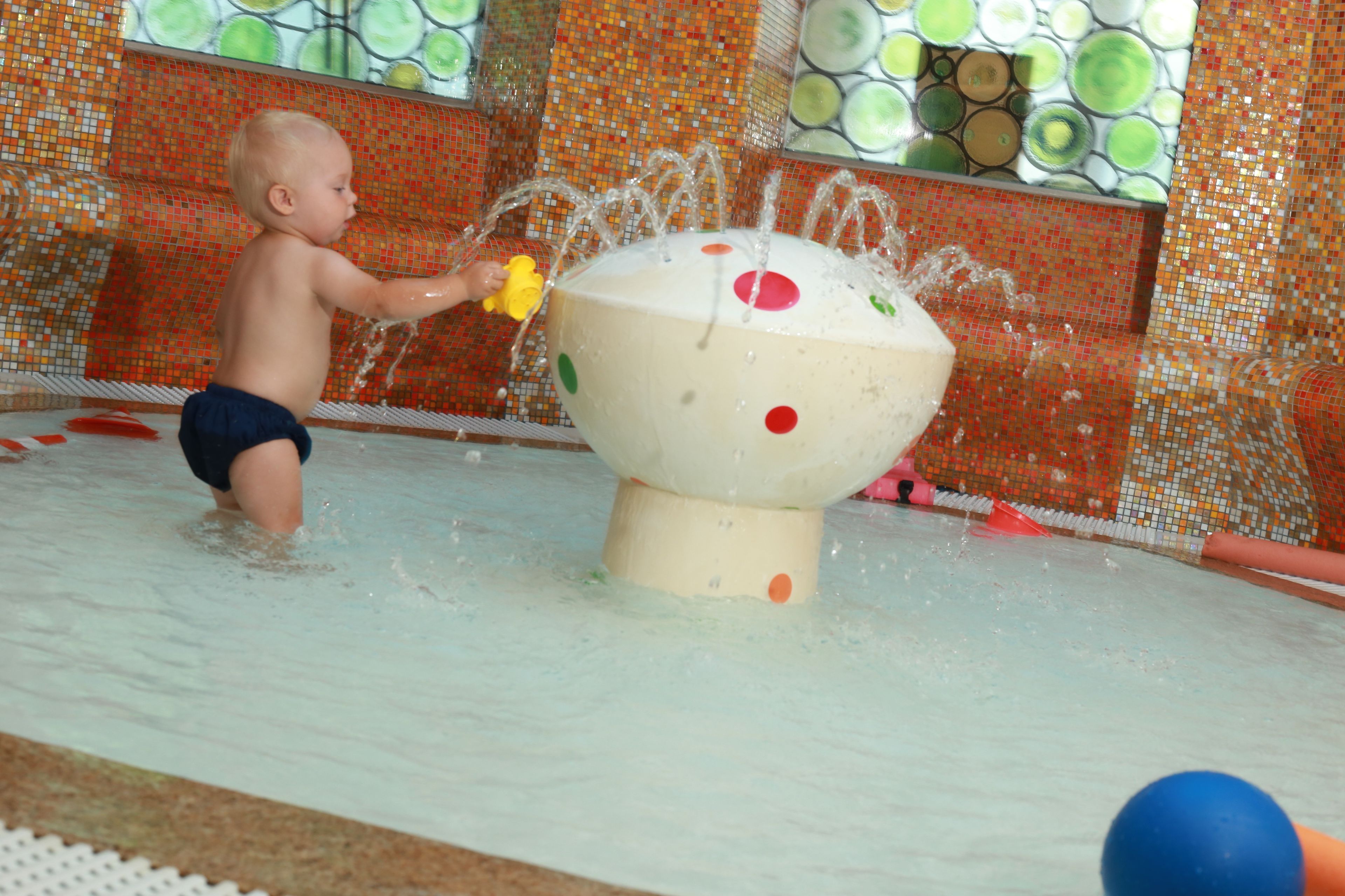 Ein Kind spielt mit Wasser in einem Poolbereich mit einer farbenfrohen Mosaikwand.