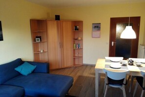 Ein Zimmer mit einem blauen Sofa, einem Holzschrank, einem Esstisch mit Stühlen und einer Pendelleuchte.