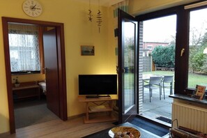 A living room with a television on a stand, a clock on the wall, and a sliding glass door leading to an outdoor patio. - Holiday apartment near the beach in Duhnen (Cuxhaven)