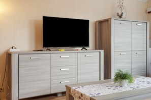 A modern living room with a flat-screen TV on a wooden cabinet, a small table with a plant, and a cabinet with multiple drawers. - Bungalow 'Haus S 41 Paulchen' with Private Terrace, Private Garden and Wi-Fi (Röbel/Müritz)