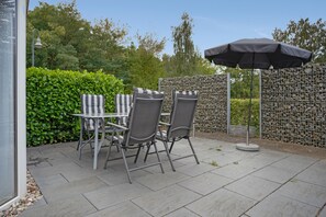 An outdoor patio with a table and chairs, a black umbrella, and a stone wall. - Bungalow 'Haus S 41 Paulchen' with Private Terrace, Private Garden and Wi-Fi (Röbel/Müritz)