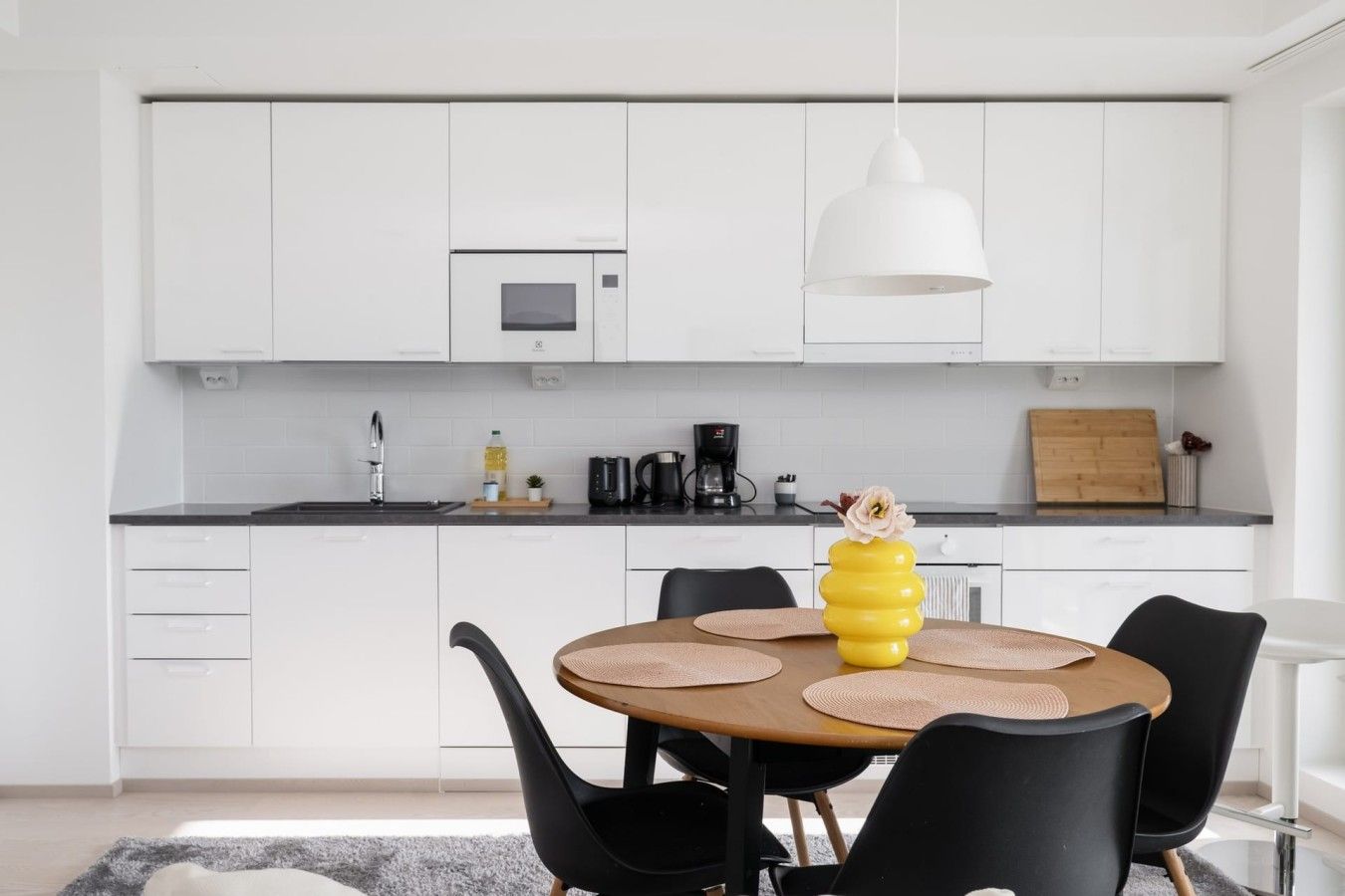 Una cocina moderna con armarios blancos, una mesa redonda de comedor con sillas negras y un jarrón amarillo con una flor.